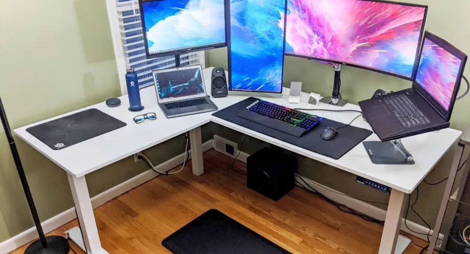 An L-shaped standing desk with a multi-monitor setup including two vertical monitors and two laptops, creating an ergonomic home office or gaming station.