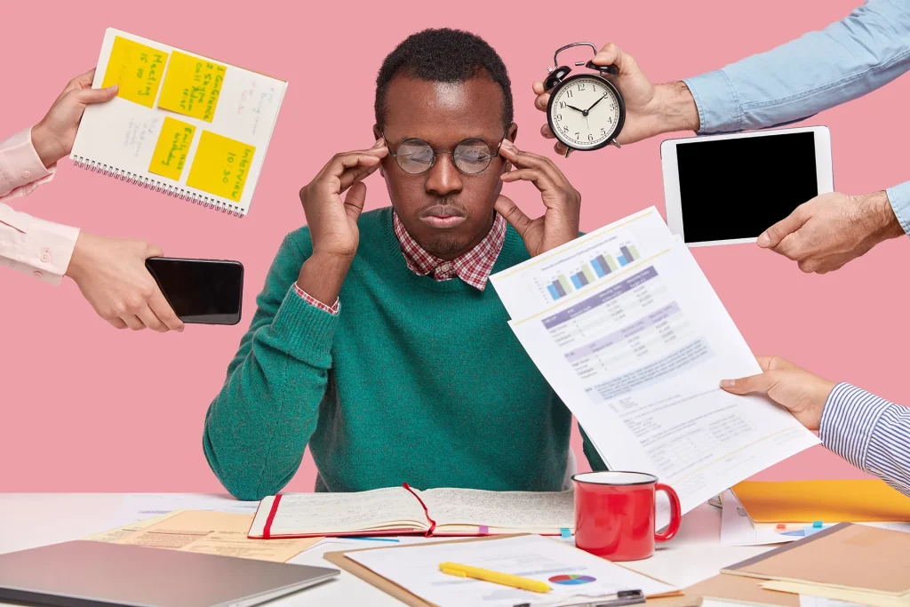 An overwhelmed man with glasses holding his head, surrounded by multiple hands offering documents, a phone, a tablet, and a clock, symbolizing stress from multitasking and tight deadlines.