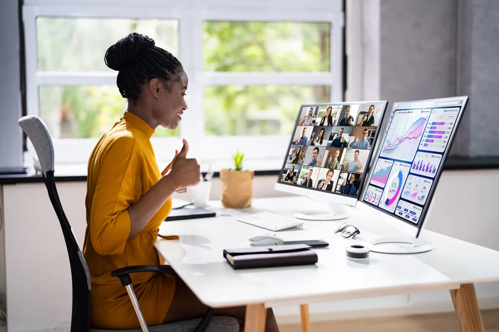 An African American woman in a yellow top giving a thumbs up during a video conference on one of her dual monitors, with the other monitor displaying business analytics, indicating a productive remote work or virtual meeting setup.