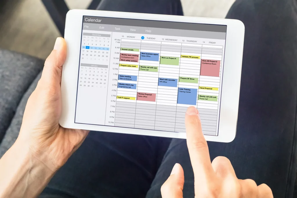 A close-up of a person's hands holding a white tablet displaying a detailed digital calendar application with color-coded events and appointments, being used for scheduling and planning.
