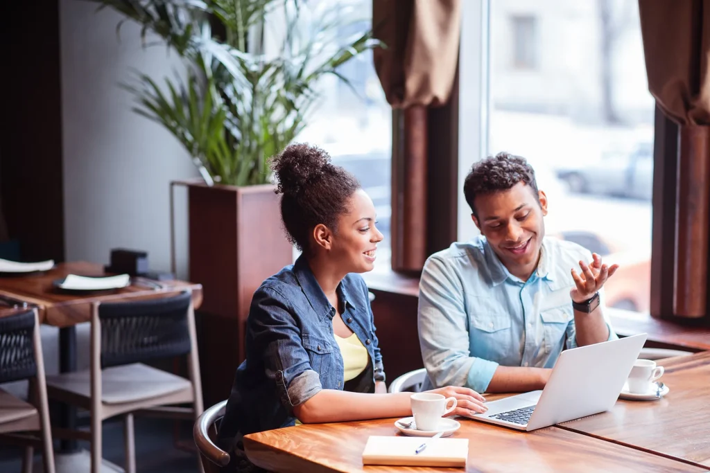 Two black people, a woman and a man, are sitting at a table in a cafe, having coffee and looking at a laptop.