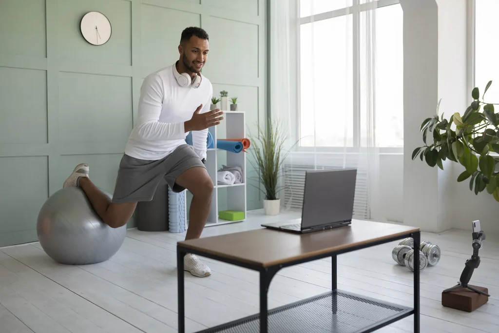 A man in athletic wear performing a lunge exercise with one foot on a Swiss ball, looking at a laptop, suggesting an online workout or home fitness routine.