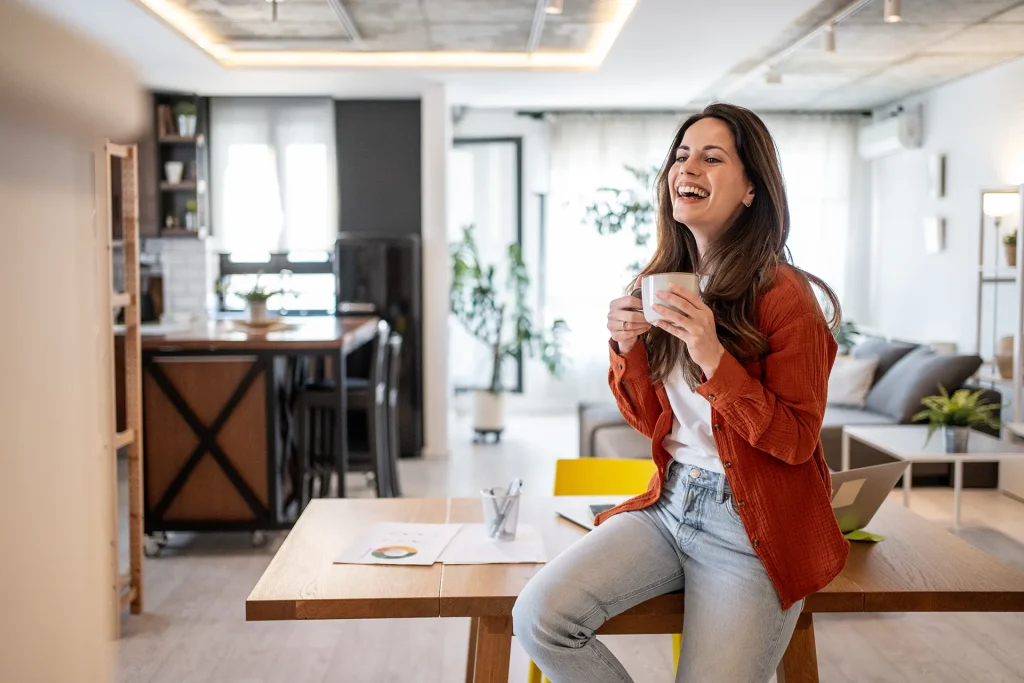 A cheerful young woman sitting on a desk with a laptop, holding a coffee cup and laughing, in a bright and comfortable home office or living room setting.