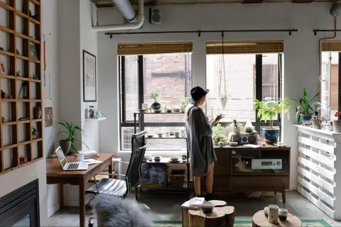 A stylish studio apartment or home office with a person tending to plants near large windows, featuring a workspace with a laptop, a wooden bookshelf, and rustic furniture, embodying a creative and cozy atmosphere.