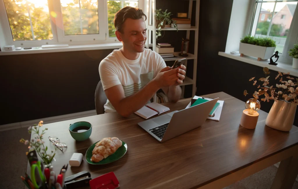 Man smiling while using his phone at his work desk with coffee and bread