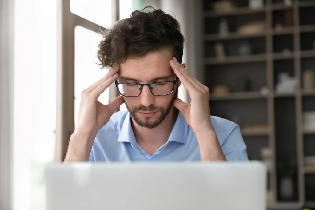 A man with glasses squinting and holding his temples in discomfort while looking at a laptop screen, suggesting a headache, eye strain, or digital fatigue.