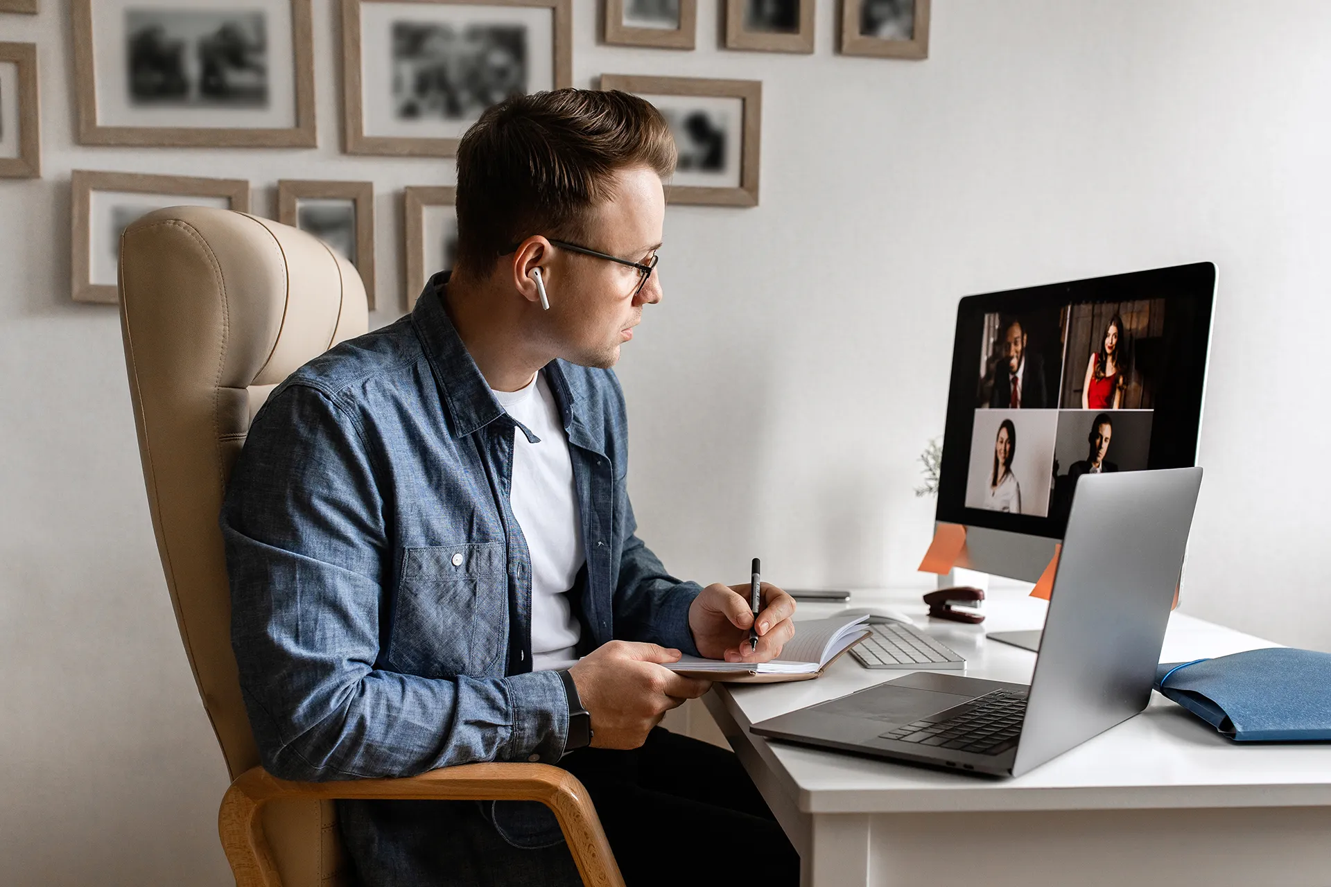 A young man with glasses and AirPods sits in an office chair, taking notes in a notebook while participating in a video conference call on his computer monitor. A laptop is also on the desk.