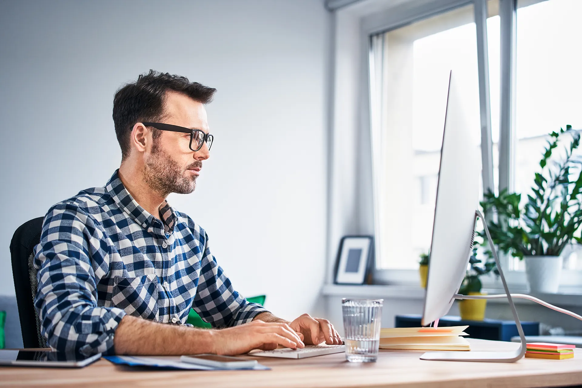 Man with glasses wearing a plaid shirt, focused while working on a desktop computer at a wooden desk in a bright home office setting.