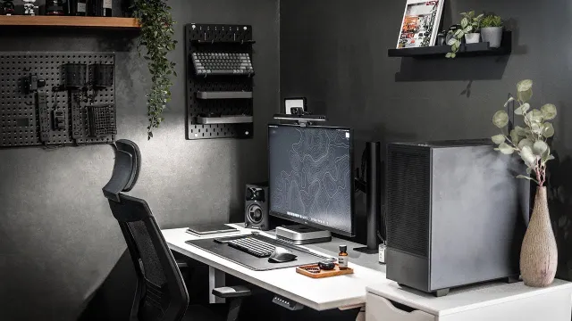 A modern, minimalist home office or gaming setup with dark walls, featuring a white desk, an ergonomic chair, a large monitor, a sleek PC case, pegboards for organization, and subtle greenery.