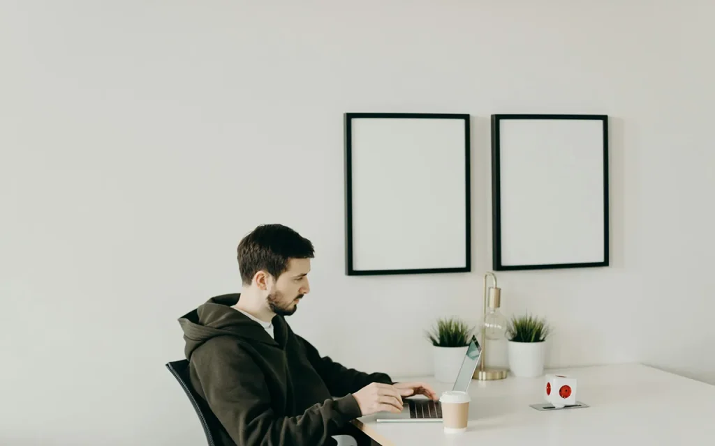 A man with a beard wearing a dark hoodie, working on a laptop at a minimalist white desk with two framed blank pictures, small potted plants, and a coffee cup, in a bright, modern home office.