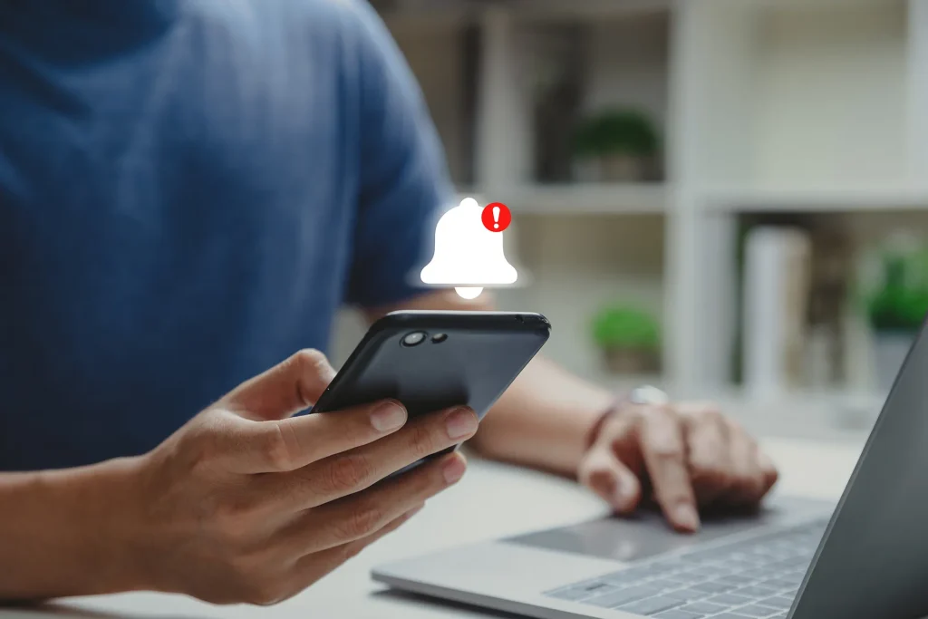 Close-up of a person holding a smartphone with a prominent white notification bell icon and a red exclamation mark, indicating a new alert or message.