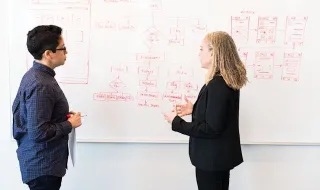 Two professionals, one man and one woman, discussing a complex diagram or flowchart drawn on a whiteboard, likely during a brainstorming session, interview, or strategic meeting.