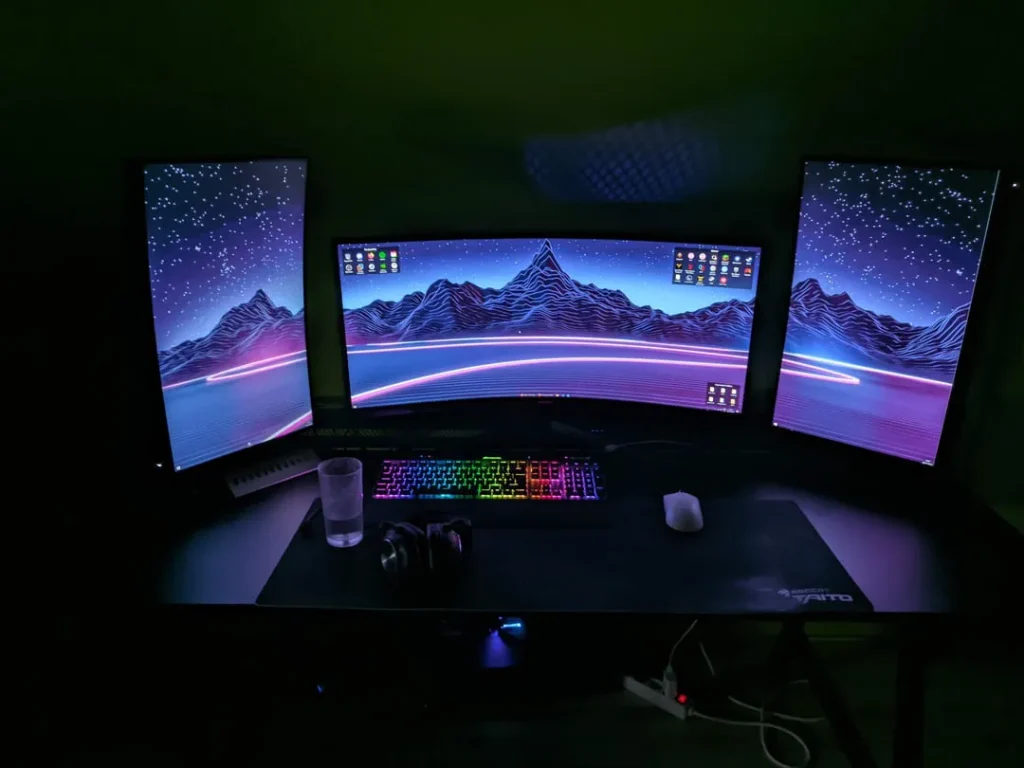 An immersive triple monitor gaming setup featuring a central curved ultrawide monitor flanked by two vertical flat monitors, all displaying a vibrant synthwave-style landscape wallpaper, with an RGB keyboard and mouse on a large desk mat.