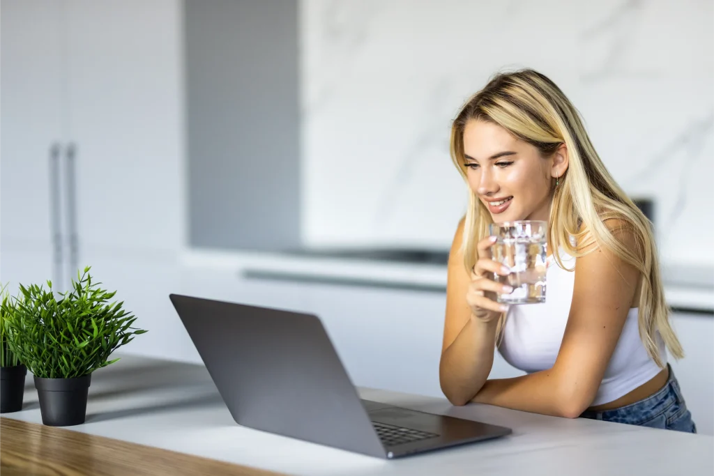 Woman using a laptop while drinking a glass of water at home