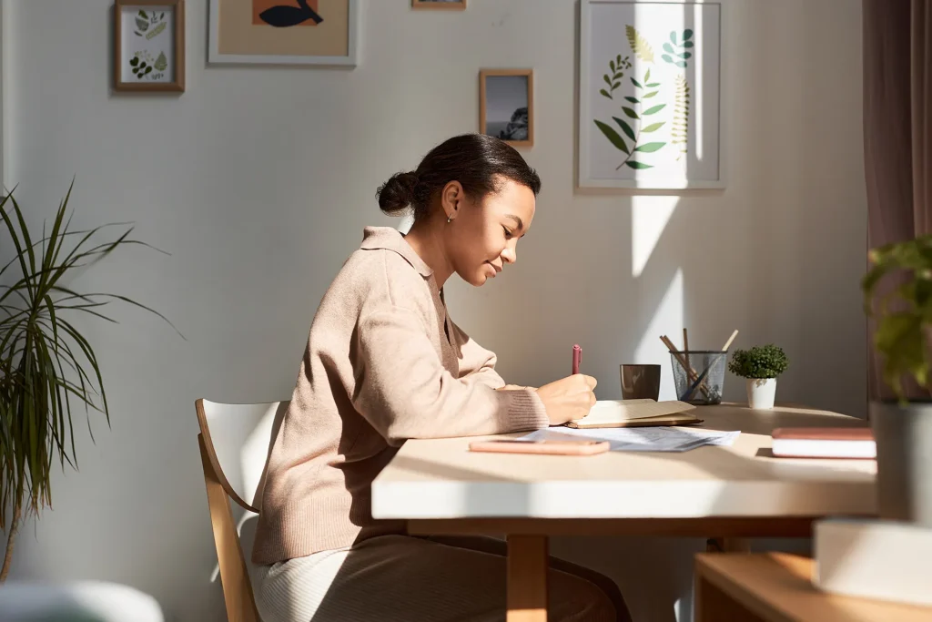 Side view of a young woman with dark hair in a bun, wearing a beige top, writing notes in a notebook at a sunlit desk with a pen, phone, and potted plant.