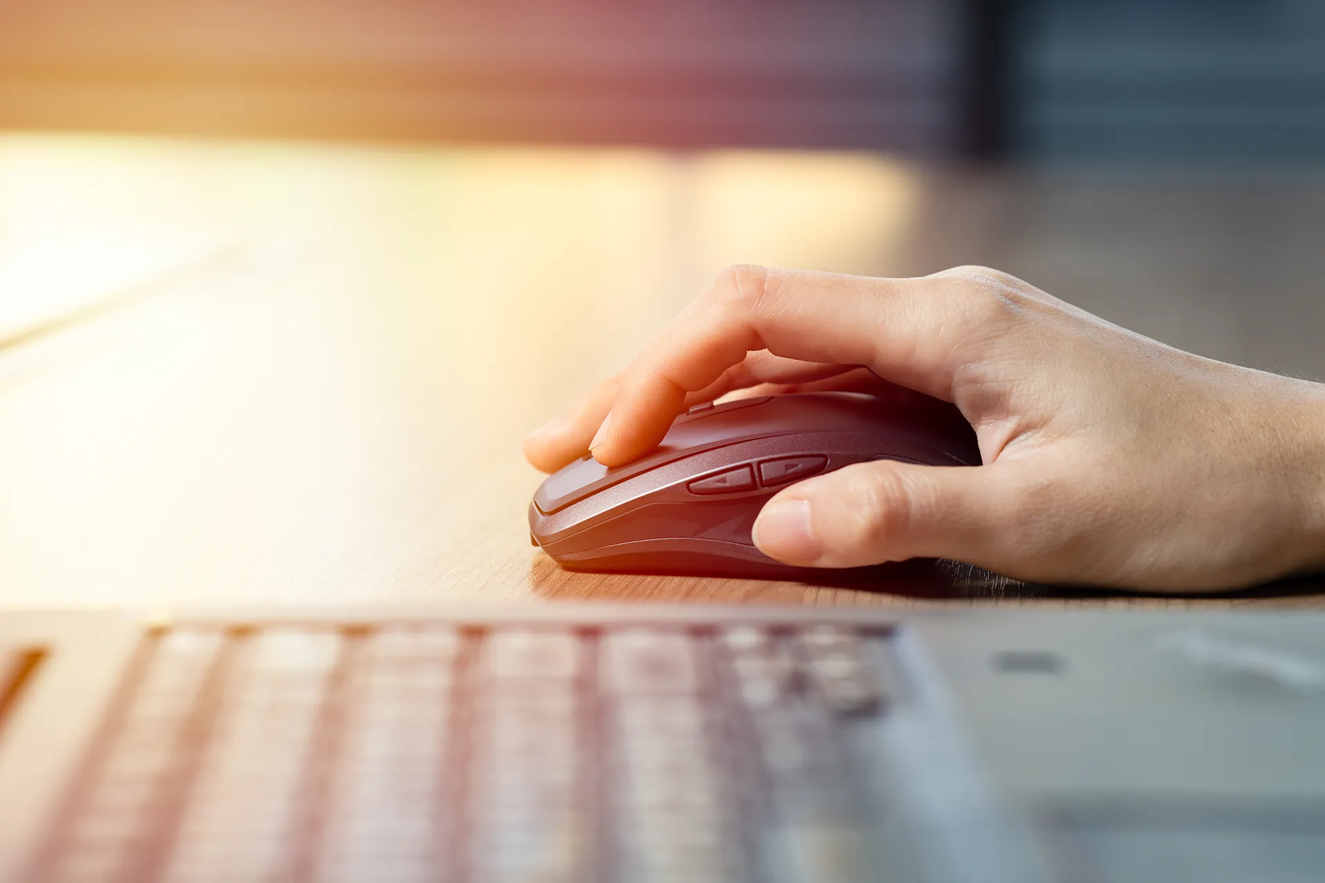 A close-up side view of a person's hand using a dark gray wireless computer mouse on a light wooden desk, with a laptop keyboard in the foreground.