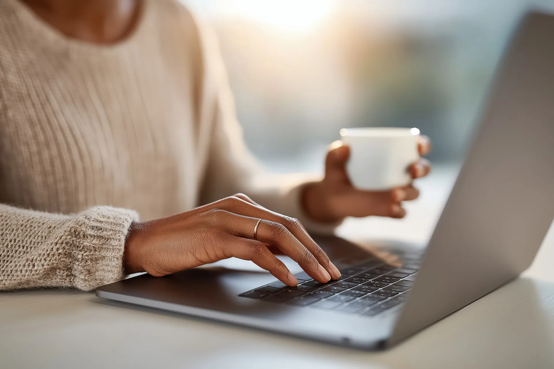 A person's hands typing on a laptop keyboard while holding a cup of coffee