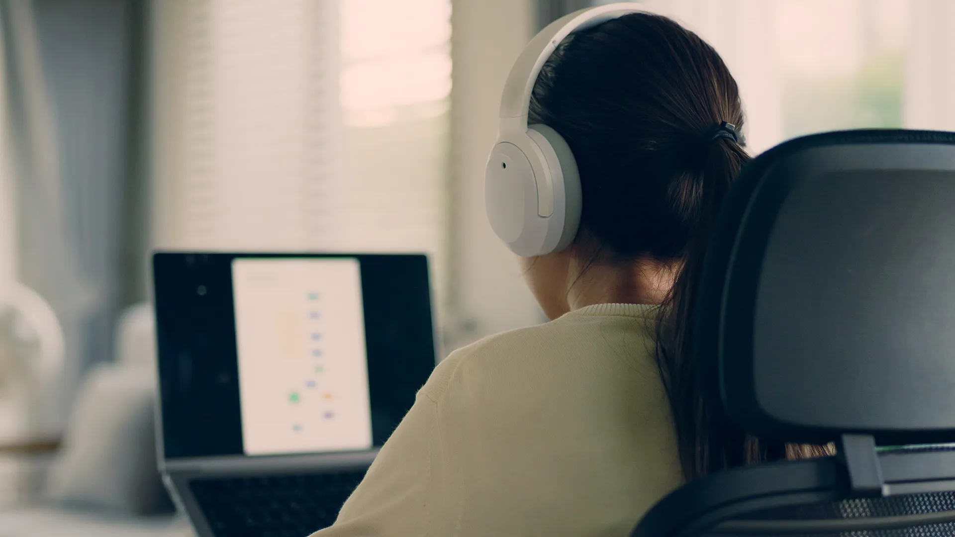 person is seen from behind, sitting in an office chair and wearing white noise cancelling headphones while looking at a laptop screen.