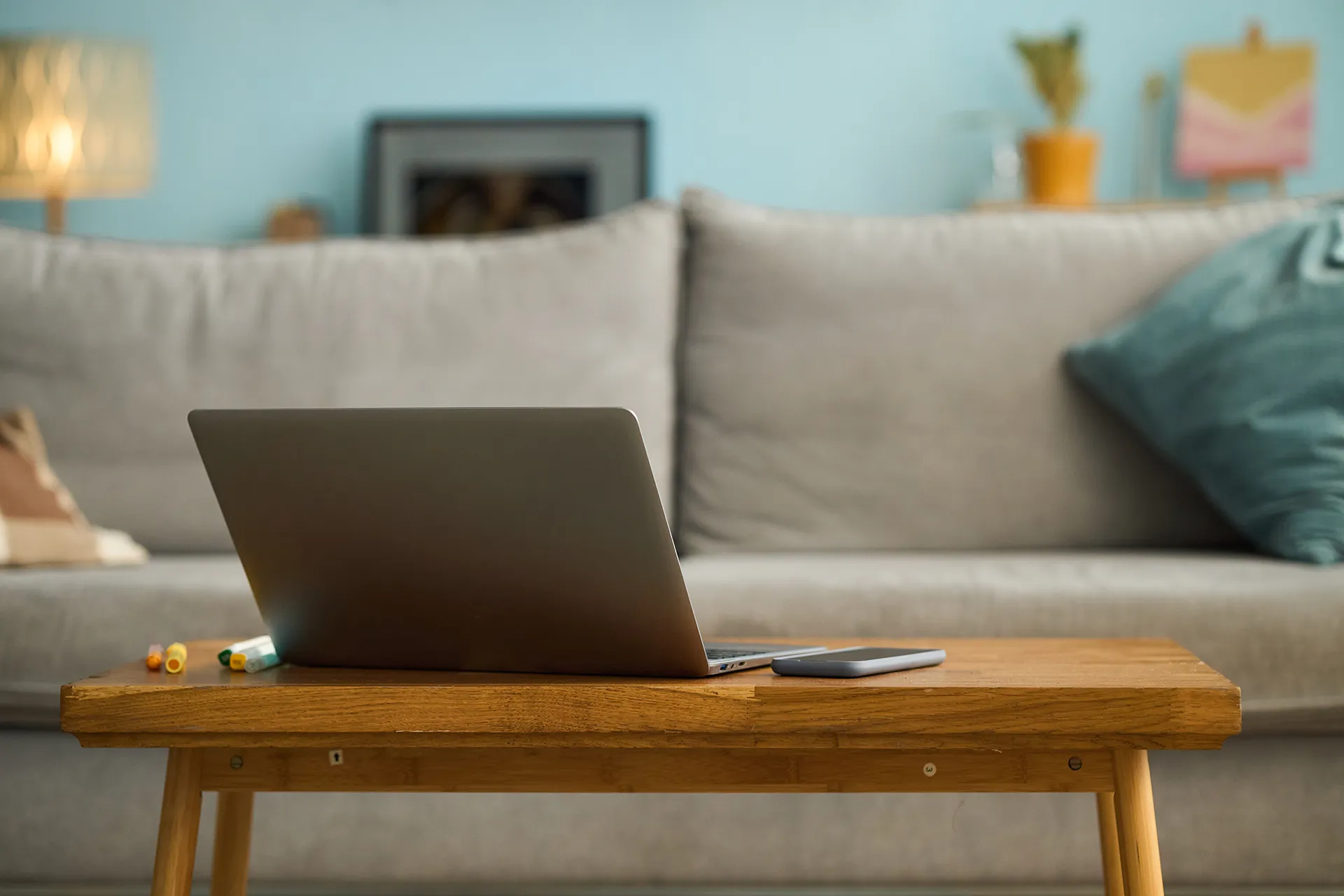 A laptop and smartphone sit on a wooden coffee table in a living room with a light gray couch and blue throw pillow in the background.