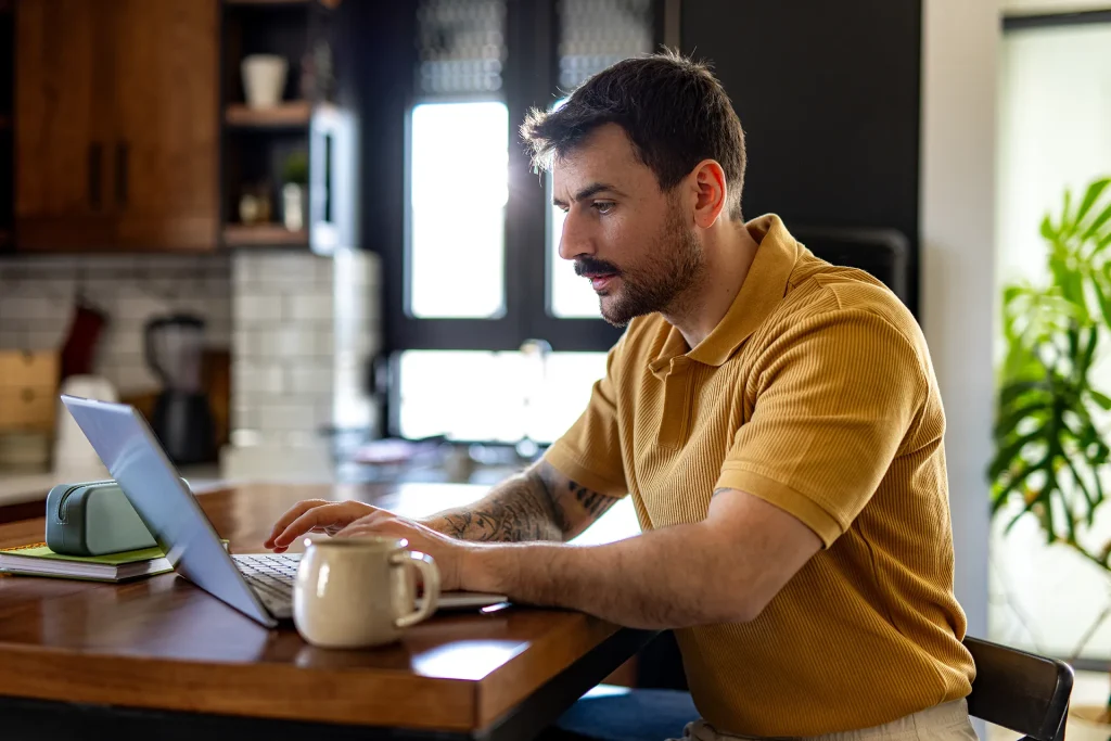 A focused man with tattoos on his arms, wearing a ribbed yellow polo shirt, working on a laptop at his kitchen counter.