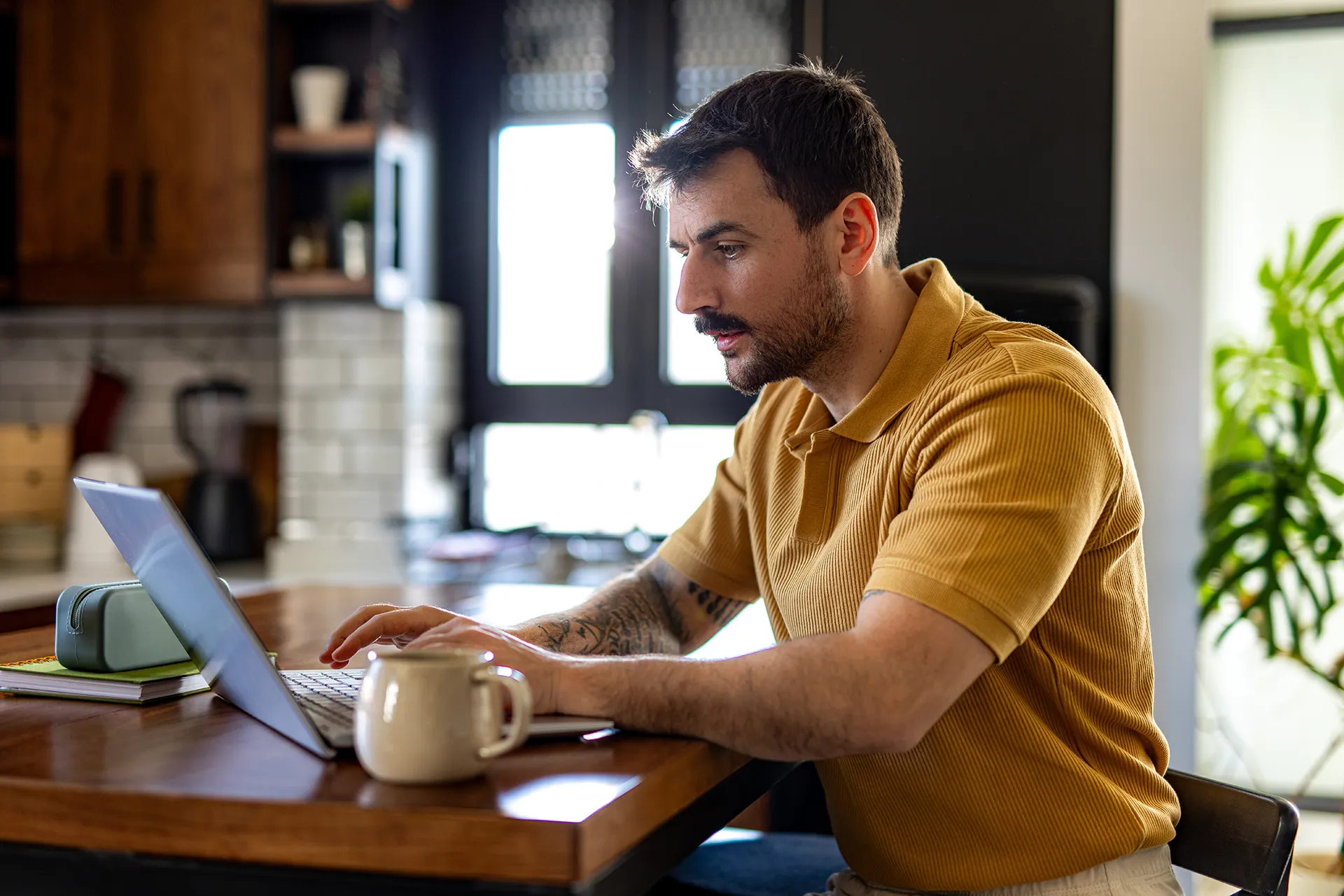 A focused man with tattoos on his arms, wearing a ribbed yellow polo shirt, working on a laptop at his kitchen counter.