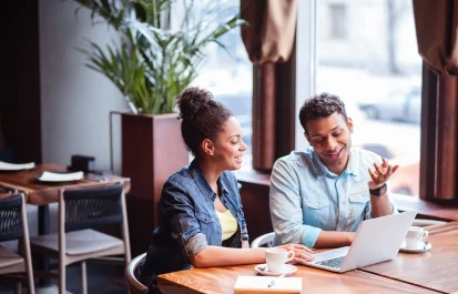 Two black people, a woman and a man, are sitting at a table in a cafe, having coffee and looking at a laptop.