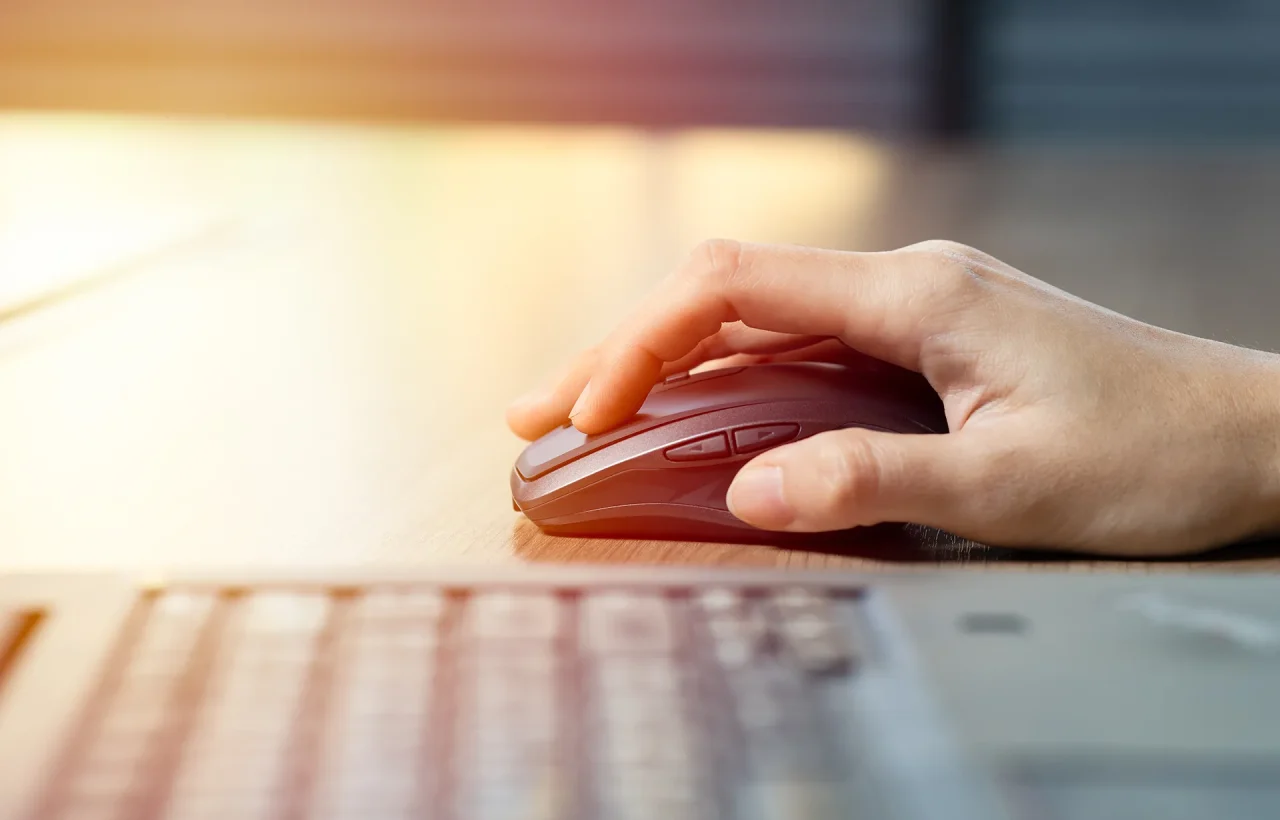 A close-up side view of a person's hand using a dark gray wireless computer mouse on a light wooden desk, with a laptop keyboard in the foreground.