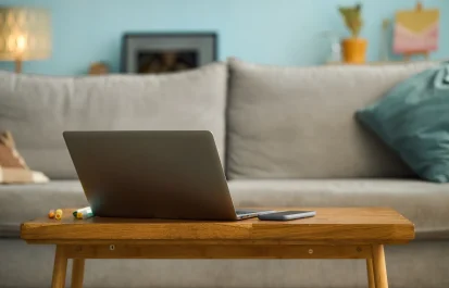 A laptop and smartphone sit on a wooden coffee table in a living room with a light gray couch and blue throw pillow in the background.