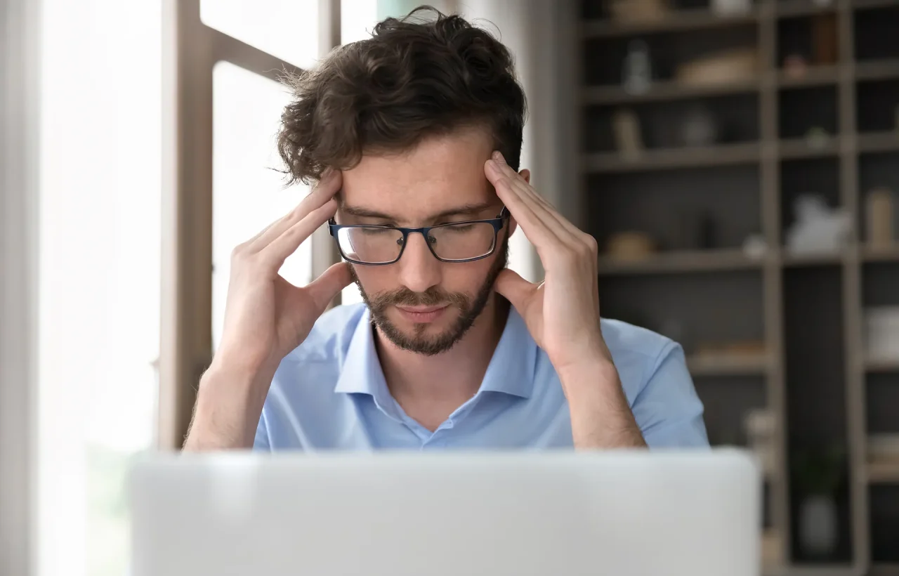 A man with glasses squinting and holding his temples in discomfort while looking at a laptop screen, suggesting a headache, eye strain, or digital fatigue.