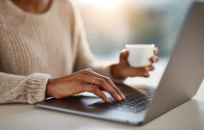 A person's hands typing on a laptop keyboard while holding a cup of coffee