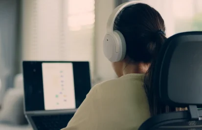 person is seen from behind, sitting in an office chair and wearing white noise cancelling headphones while looking at a laptop screen.